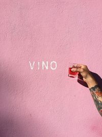 Cropped tattooed hand of person holding red drink against pink wall