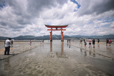 People by torii gate in river against cloudy sky