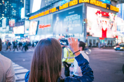 Rear view of woman photographing with mobile phone while standing against building at times square
