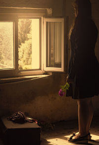 Young woman holding flower looking through window while standing in room
