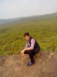 Full length side view portrait of young man crouching on mountain against sky