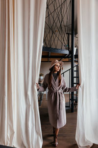 A girl in a beige hat and a beige coat stands indoors and pushes the white curtain with her hands