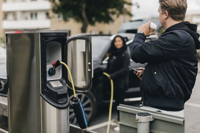 Man drinking coffee while standing at electric car charging station