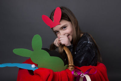 Portrait of smiling girl with costume rabbit ears sitting in basket against gray background