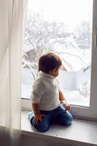 Boy in a white sweater standing at the window on the windowsill in winter