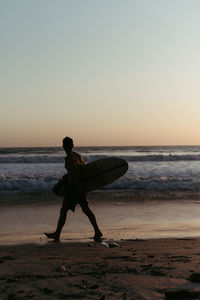Side view of anonymous man silhouette holding surfboard while walking along sandy seashore in summertime during sunset
