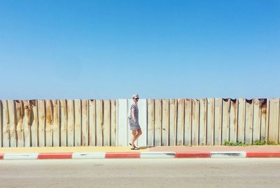 Side view of mid adult woman walking on footpath by fence against clear blue sky during sunny day