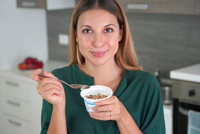 Attractive woman holds bowl of greek yogurt with granola with blurred kitchen on the background