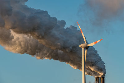 Low angle view of wind turbine by smoke emitting from chimney against blue sky