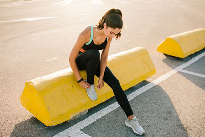 Full body of young slim female runner in black sportswear sitting on concrete border on paved street and tying laces on sneakers while preparing for jogging