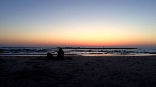 Silhouette people sitting on beach against clear sky during sunset
