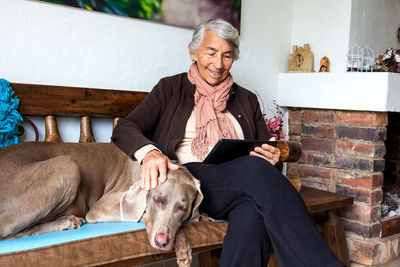 Senior woman using digital tablet with dog sitting on bench at home