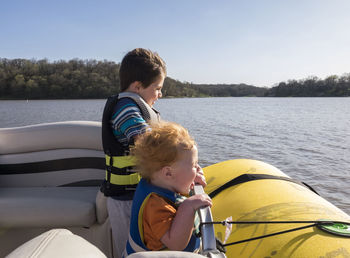 Happy brothers wearing life jackets while standing in boat on lake against clear sky during sunny day