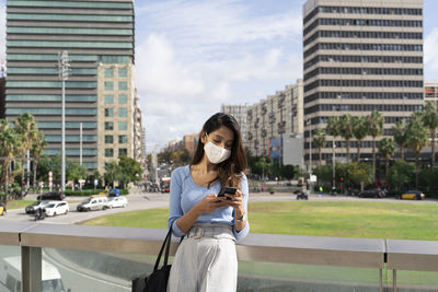 Businesswoman in protective face mask using smart phone while leaning on railing during coronavirus outbreak
