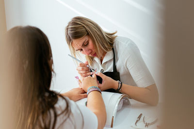 Concentrated female manicurist using brush while applying acrylic nail powder on tips of anonymous client in daylight in beauty salon