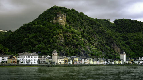 Town at riverbank by tree mountains against cloudy sky