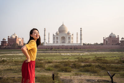Side view of delighted asian female tourist standing near historic mausoleum while sightseeing in india and looking at camera