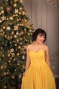 Portrait of smiling beautiful woman wearing yellow dress while standing by christmas tree at home