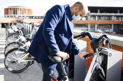 Man wearing protective face mask unlocking bicycle through smart phone on sunny day