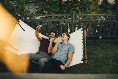 High angle view of smiling friends taking selfie on smart phone while resting in hammock at yard