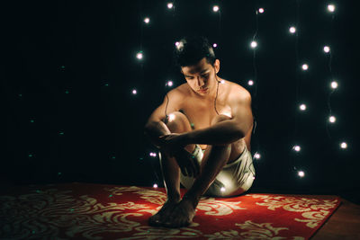 Depressed shirtless man amidst illuminated fairy lights against black background