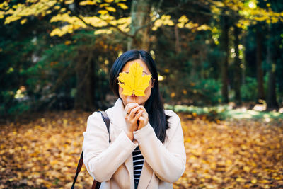 Woman hiding face with dry yellow leaf against trees in forest during autumn