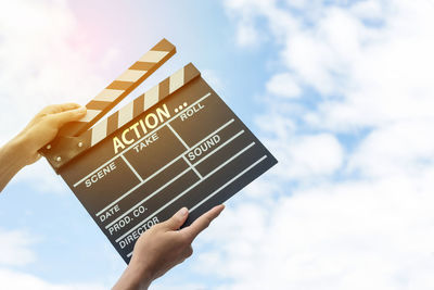 Low angle view of hands holding film slate with text against sky