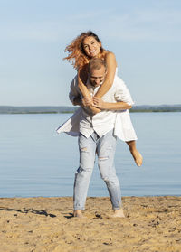 Happy smiling caucasian man carries laughing latin woman on back, blue sky,water on background