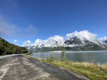 Scenic view of fjord against mountain covered in clouds with clear sky above. road in front.