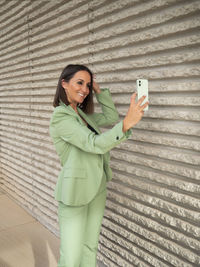 Young woman using mobile phone while standing against wall