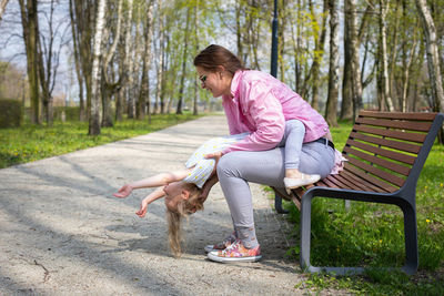 A mother and daughter play gymnastics while sitting on a bench in a city park.