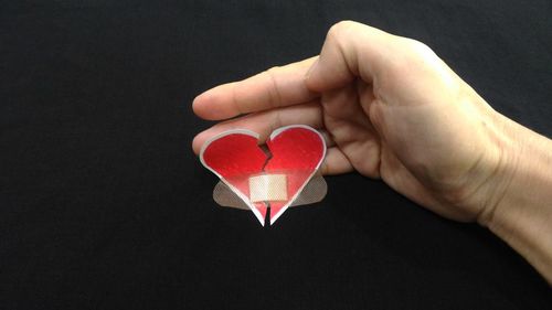 Cropped hand holding torn heart shape paper with bandage against black background