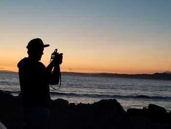 Silhouette man photographing sea against sky during sunset