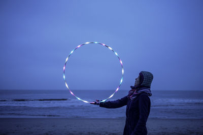 Side view of woman holding illuminated plastic hoop at beach against clear sky at dusk