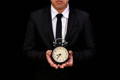 Midsection of businessman holding alarm clock against black background