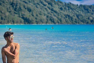 Shirtless man wearing sunglasses while standing at beach against mountain