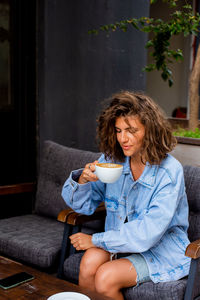 Young charming woman with curly hairstyle holding big mug, having fresh cappuccino 