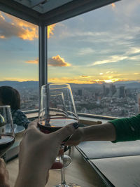 Hand holding glass of cityscape against sky during sunset