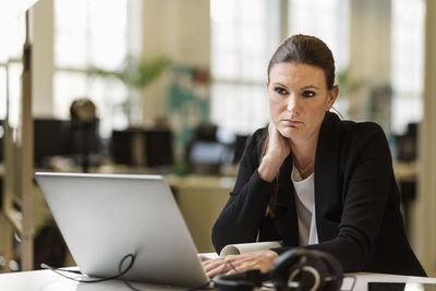 Thoughtful businesswoman looking away while using laptop at desk in creative office