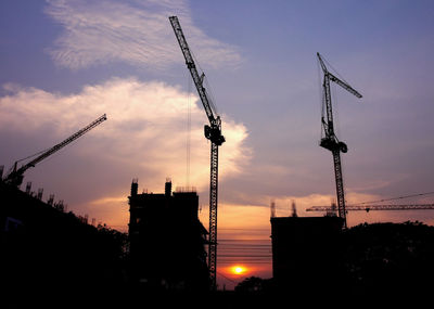 Low angle view of silhouette crane by building against sky during sunset