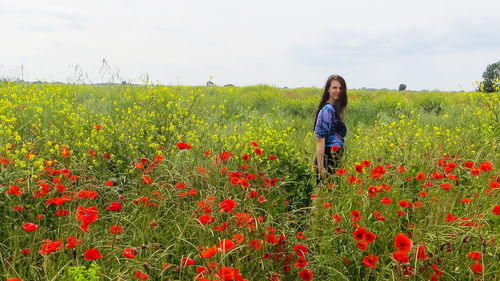 Portrait of woman standing by poppy flowers on field against sky
