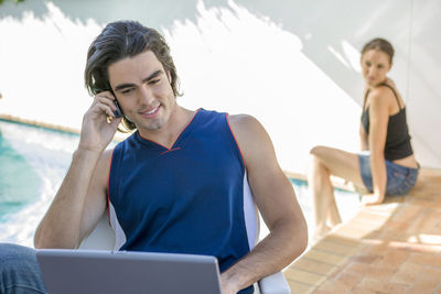Smiling man talking on mobile phone while using laptop with girlfriend in background