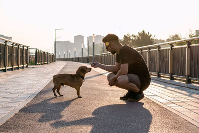 Young handsome man is feeding his dog from hand in the morning on empty street of city.