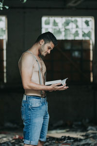 Side view of shirtless young man reading book standing at home