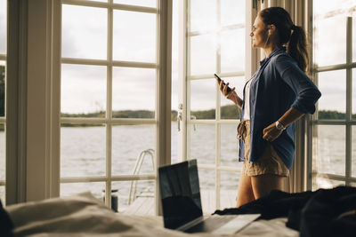 Thoughtful mature woman listening music through headphones on smart phone at holiday villa