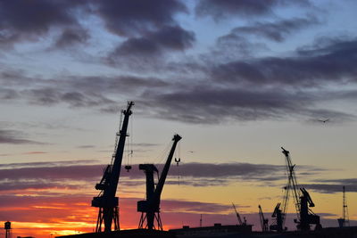 Silhouette cranes at harbor against sky during sunset