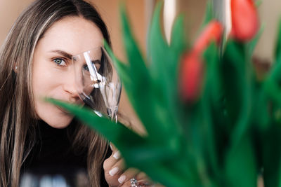 Woman against background of blurred bouquet of flowers, covering one eye with wine glass