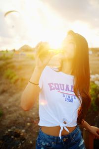 Young woman drinking drink while standing against sky during sunset