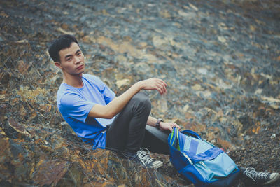 Portrait of young man holding backpack while sitting on rock