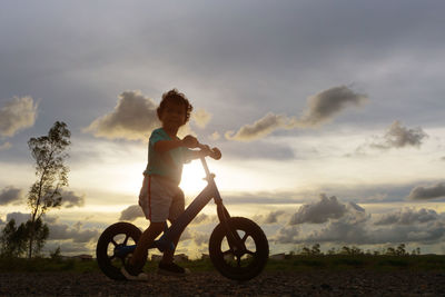 Boy riding bicycle against cloudy sky at sunset